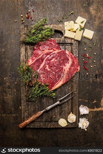 Raw beef steak with thyme,butter and meat fork on dark rustic cutting board, top view