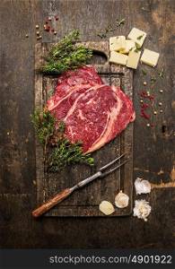 Raw beef steak with thyme,butter and meat fork on dark rustic cutting board, top view