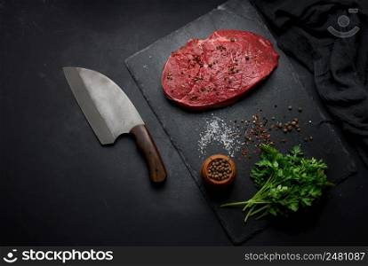 Raw beef tenderloin lies on a cutting board and spices for cooking on a black table, top view