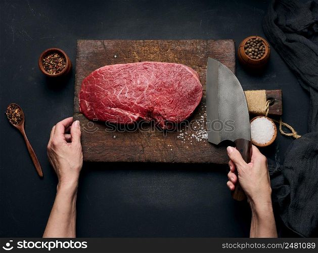 Raw beef tenderloin lies on a wooden cutting board and spices for cooking on a black table, top view. A woman&rsquo;s hand holds a large kitchen knife in her hand