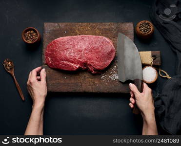 Raw beef tenderloin lies on a wooden cutting board and spices for cooking on a black table, top view. A woman&rsquo;s hand holds a large kitchen knife in her hand