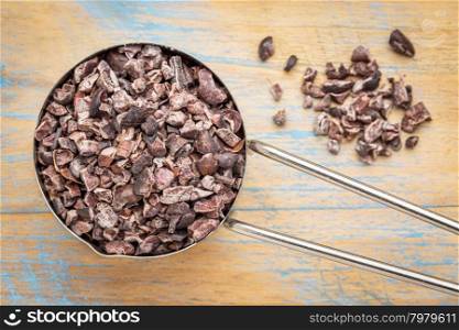 Raw cacao nibs in a metal measuring scoop against wooden background