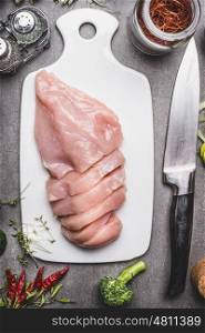 Raw chicken breast fillets on white cutting board with kitchen knife and ingredients for cooking, top view, close up