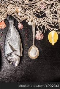 raw dorado fish with lemon, peppercorn, spoon of salt, and fishing net on black stone background, top view, vertical