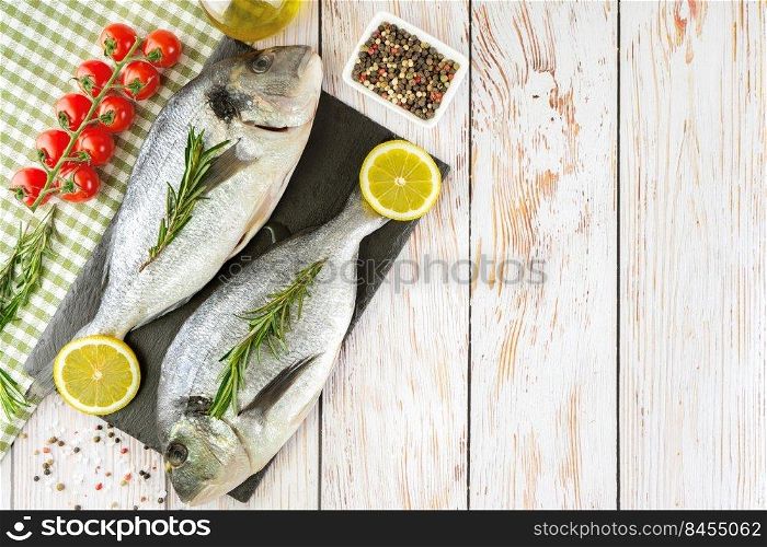 Raw fish dorado on black cutting board on white wooden background with spices, tomato, rosemary, olive oil and lemon. Top view, flat lay with copy space for text. Fresh fish dorado on white wooden background with ingredients for cooking