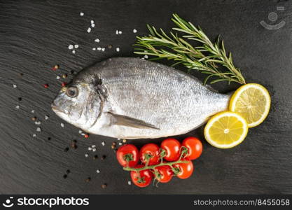 Raw fish dorado on black slate background with spices, tomato, rosemary, olive oil and lemon. Top view, flat lay with copy space for text. Fresh fish dorado on black slate background with ingredients for cooking