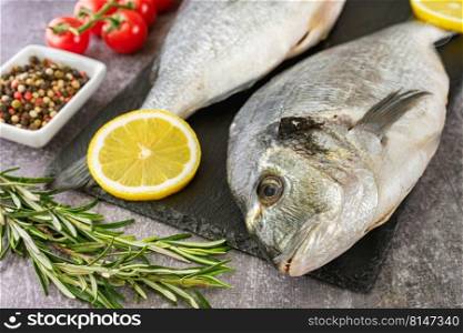 Raw fish dorado on black slate cutting board and grey concrete background with spices, tomato, rosemary, olive oil and lemon. Fresh fish dorado on black slate cutting board and grey concrete background with ingredients for cooking