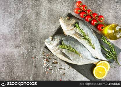 Raw fish dorado on black slate cutting board and grey concrete background with spices, tomato, rosemary, olive oil and lemon. Top view, flat lay with copy space for text. Fresh fish dorado on black slate cutting board and grey concrete background with ingredients for cooking