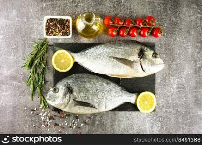 Raw fish dorado on black slate cutting board and grey concrete background with sπces, tomato, rosemary, olive oil and≤mon. Top view, flat lay with©space for text. Fresh fish dorado on black slate cutting board and grey concrete background with ingredients for cooking