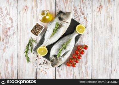 Raw fish dorado on black slate cutting board and white wooden background with spices, tomato, rosemary, olive oil and lemon. Top view, flat lay with copy space for text. Fresh fish dorado on black slate cutting board and white wooden background with ingredients for cooking