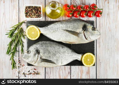 Raw fish dorado on black slate cutting board and white wooden background with spices, tomato, rosemary, olive oil and lemon. Top view, flat lay with copy space for text. Fresh fish dorado on black slate cutting board and white wooden background with ingredients for cooking