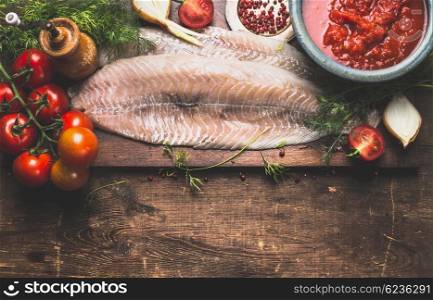 Raw fish fillet with frish herbs, tomatoes and sauce on dark rustic cutting board on dark aged wooden background, top view, border. Pollack fish cooking