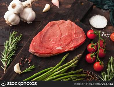 Raw fresh beef braising steak on chopping board with garlic, asparagus and tomatoes with salt and pepper on wooden table background. Macro