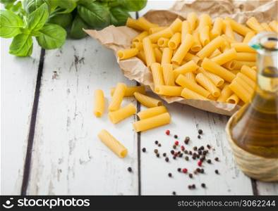 Raw fresh homemade penne pasta in brown paper with basil and oil on white wooden table background.