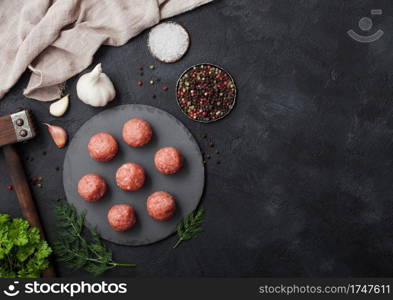 Raw fresh minced beef meatballs on round board with pepper, salt and garlic on black background with parsley and wooden hammer