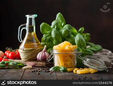 Raw fresh penne pasta in glass jar with oil and garlic, basil plant on wooden table background. Macro