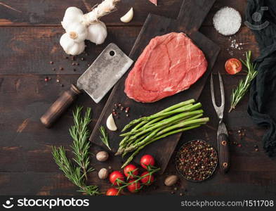 Raw fresh slice of beef braising steak on chopping board with garlic, asparagus and tomatoes with salt and pepper with rosemary on wooden background. Top view