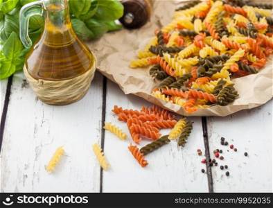 Raw homemade tricolore fusilli pasta in brown paper on white wooden table background with basil and oil.