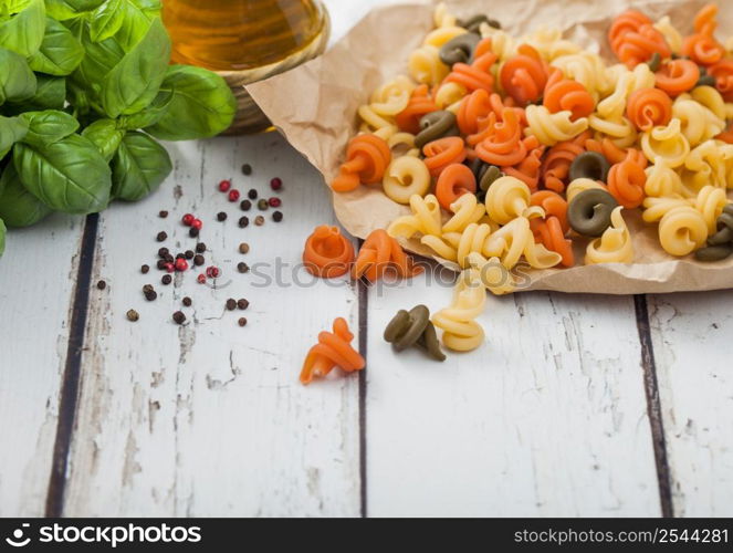 Raw homemade tricolore trottole pasta in brown paper on white wooden table background with basil and oil