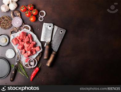 Raw lean diced casserole beef pork steak on chopping board with vintage meat hatchets on brown background. Salt and pepper with rosemary and red onion and garlic