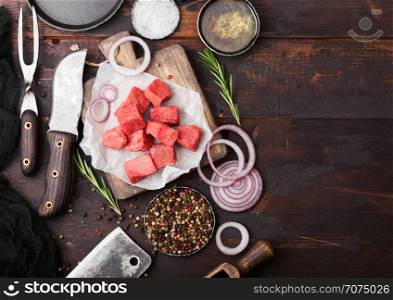 Raw lean diced casserole beef pork steak with vintage meat hatchet and knife and fork on wooden background. Salt and pepper with fresh rosemary, red onion and garlic.