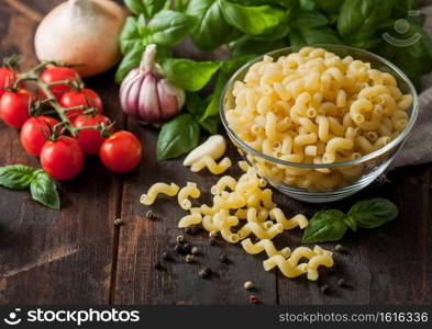 Raw maccheroni elbows pasta in glass bowl with oil and garlic, basil plant and tomatoes with pepper on wooden table. Top view