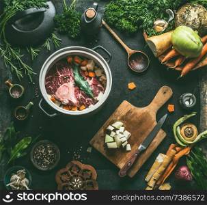 Raw meat in cooking pot on kitchen table background with vegetables , seasoning and kitchen utensils, top view. Flat lay. Meat dishes recipes. Broth, meat bone stock or soup cooking preparation