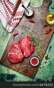Raw meat steaks on wooden cutting board with oil, herbs and spices. Rustic background, top view
