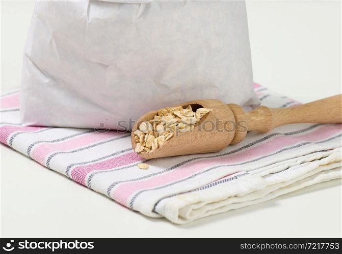 raw oatmeal in a white paper bag and a wooden spoon on a white table, breakfast porridge