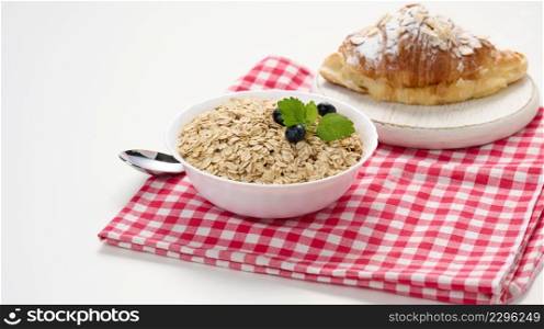 Raw oatmeal in white ceramic plate and croissant on white table, breakfast