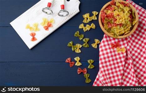 raw pasta in the form of bows in a wooden round plate and white textile napkin on a blue wooden background, top view. Mediterranean food set.