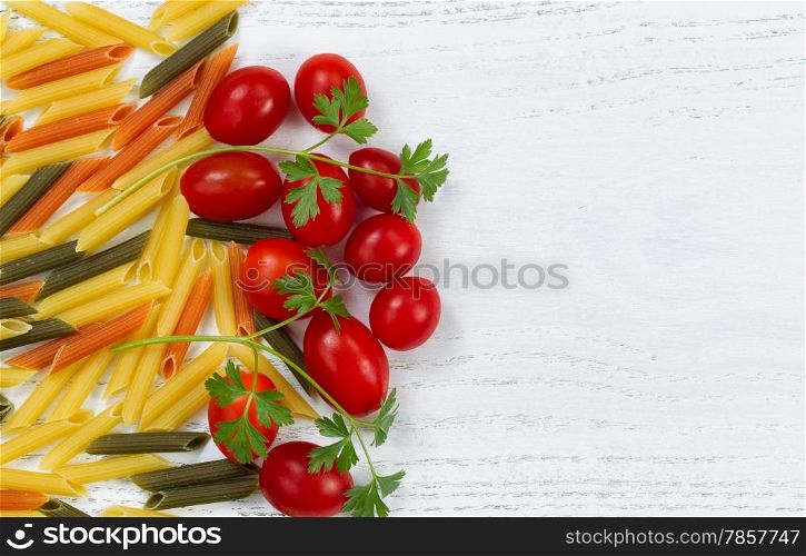 Raw pasta, of different colors, small grape tomatoes and parsley on rustic white wood. Top view angled shot.
