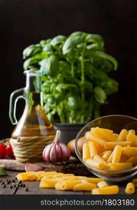 Raw penne pasta in glass bowl with oil and garlic, basil plant and tomatoes on wooden table background.