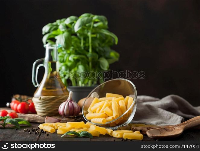Raw penne pasta in glass bowl with oil and garlic, basil plant and tomatoes with pepper and linen towel on wooden table. Space for text