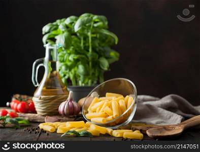 Raw penne pasta in glass bowl with oil and garlic, basil plant and tomatoes with pepper and linen towel on wooden table. Space for text