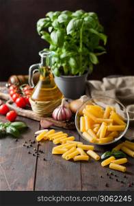 Raw penne pasta in glass bowl with oil and garlic, basil plant and tomatoes with pepper and linen towel on wooden table background.