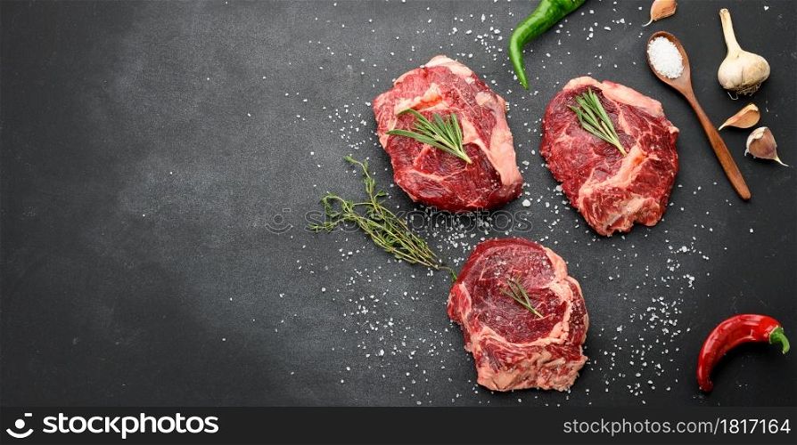 raw piece of beef ribeye with rosemary, thyme on a black table, top view, copy space