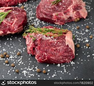 raw piece of beef ribeye with rosemary, thyme on a black table, close up