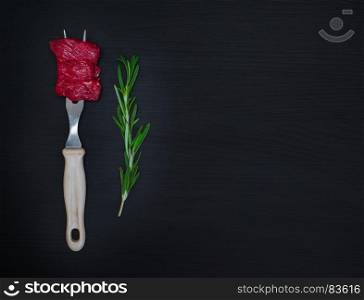 raw pieces of meat on an iron kitchen fork and a green branch of rosemary on a black wooden background, empty space on the right