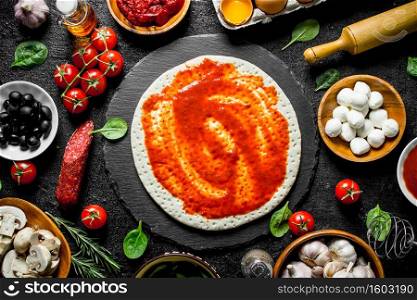 Raw pizza. Rolled out dough with tomato paste, sausage, mozzarella and olives. On black rustic background. Raw pizza. Rolled out dough with tomato paste, sausage, mozzarella and olives.