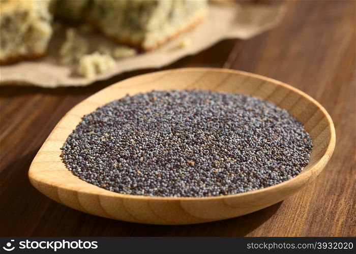 Raw poppy seeds on small wooden plate, poppy seed cake in the back, photographed with natural light (Very Shallow Depth of Field, Focus one third into the poppy seeds)