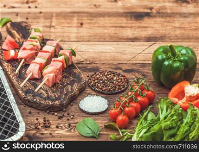 Raw pork kebab with paprika on chopping board with fresh vegetables and disposable charcoal grill on wooden background with fork and knife. Salt and pepper with lettuce and paprika.