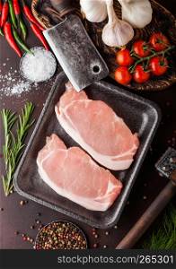 Raw pork loin chops in plastic tray with salt and pepper and vintage meat hatchets and hammer on rusty board.Red pepper, tomatoes and garlic.