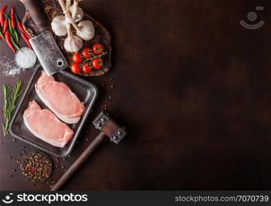 Raw pork loin chops in plastic tray with salt and pepper and vintage meat hatchets and hammer on rusty background.Red pepper, tomatoes and garlic.