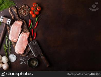 Raw pork loin chops on old chopping board with salt and pepper and vintage meat hatchets and hammer on rusty background.Red pepper, tomatoes and garlic.
