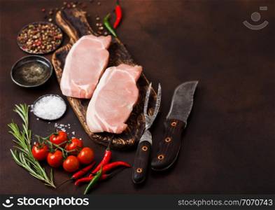 Raw pork loin chops on old vintage chopping board with knife and fork on rusty board. Salt and pepper with fresh rosemary and oil with pepper and tomatoes.
