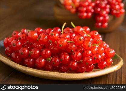 Raw red currants (lat. Ribes rubrum) on plate, photographed on dark wood with natural light (Selective Focus, Focus one third into the redcurrants)