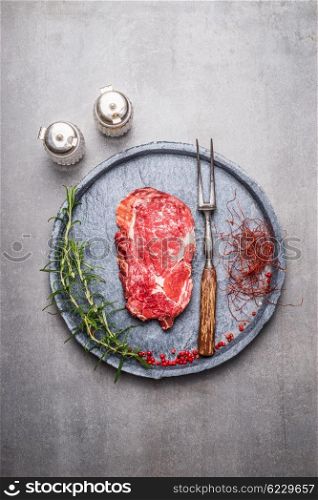 Raw Ribeye Steak with spices and meat fork on gray stone plate , top view