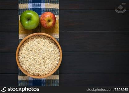 Raw rolled oats in wooden bowl with apples on cloth, photographed on dark wood with natural light