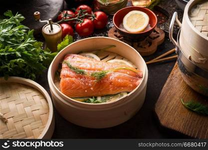 Raw salmon fillet in bamboo steamer on dark rustic kitchen table with ingredients and tools. Healthy eating and cooking. Dieting concept. Asian cuisine